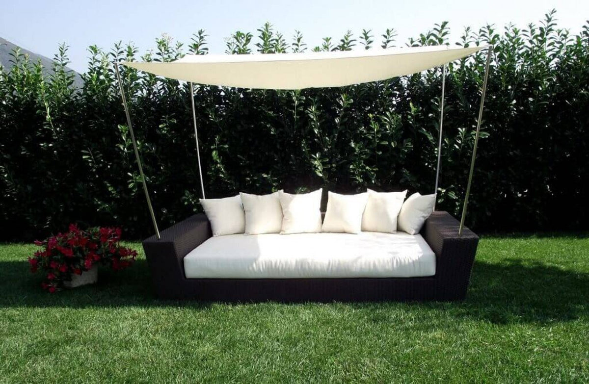 Divano SHADDY marrone in wicker con tenda ecru su prato, vista frontale tra siepi e fiori