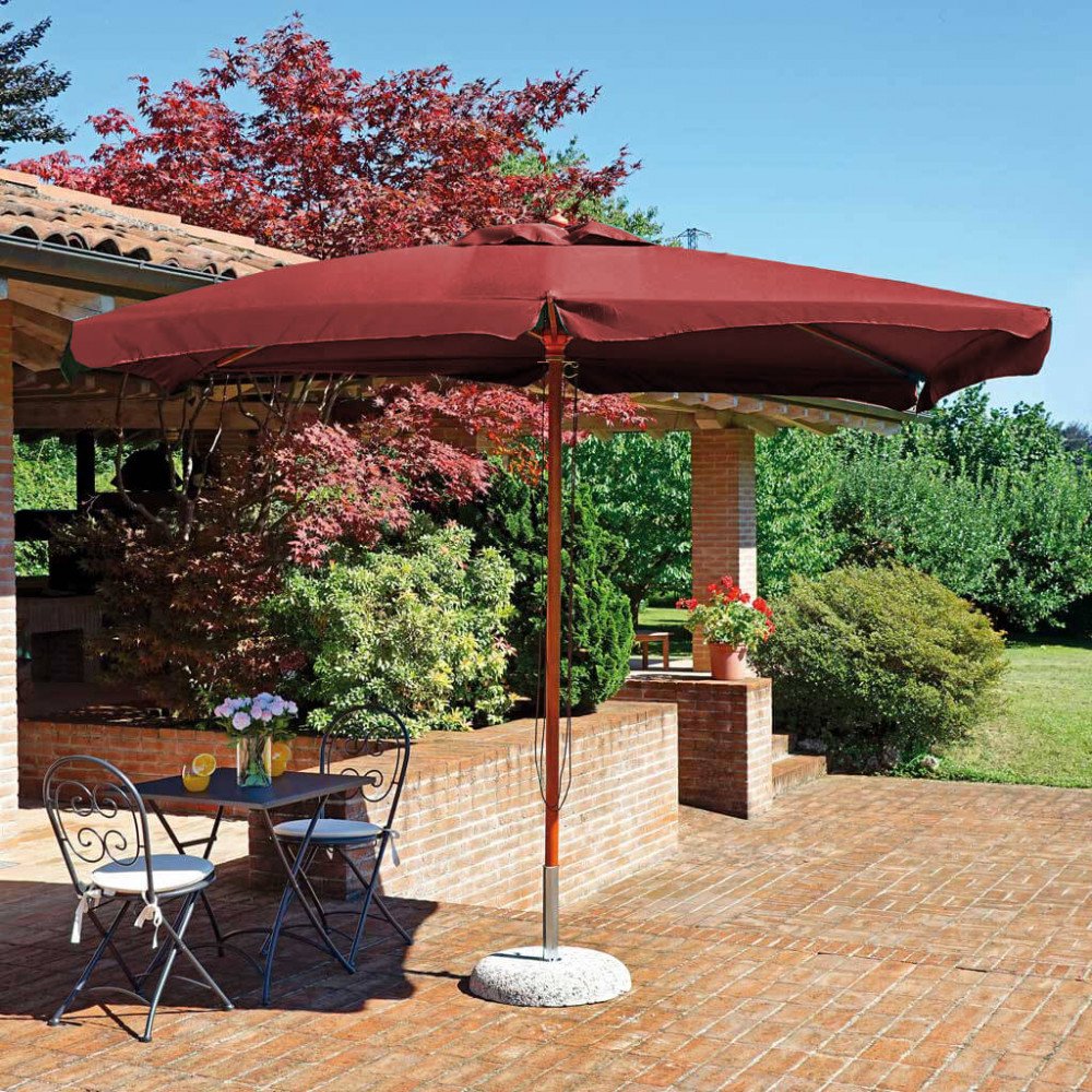 Ombrellone LENA rosso bordeaux ripreso di tre quarti, base in pietra e muretto in mattoni #colore_Rosso